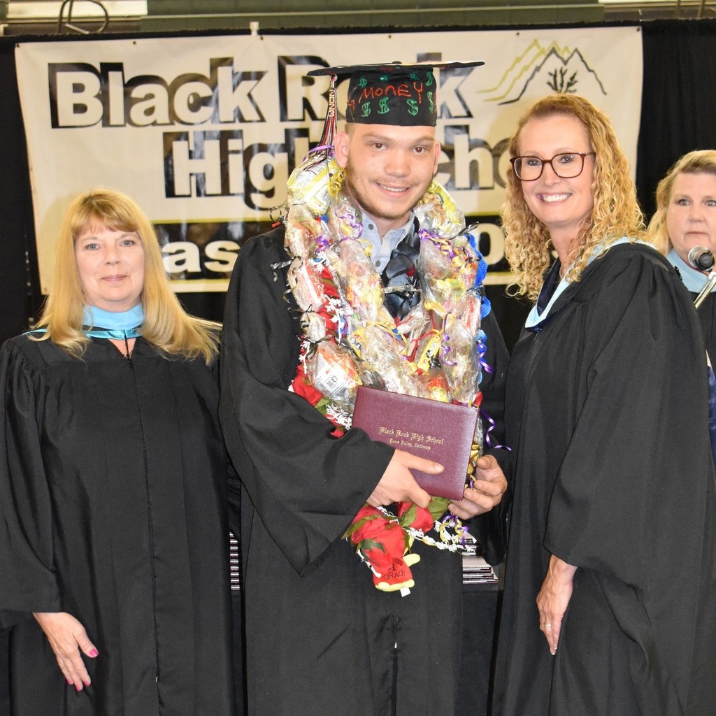 a graduate with a decorated cap decked out in honor cords, with smiling teachers