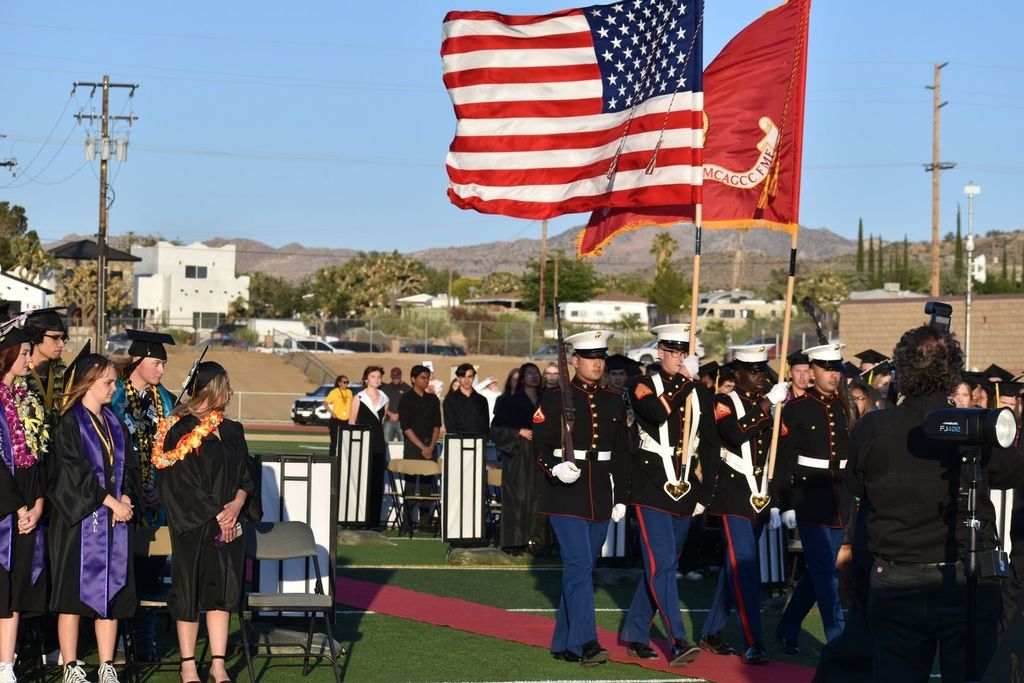 military officers in uniform carrying the American and California flags past assembled students in graduation caps